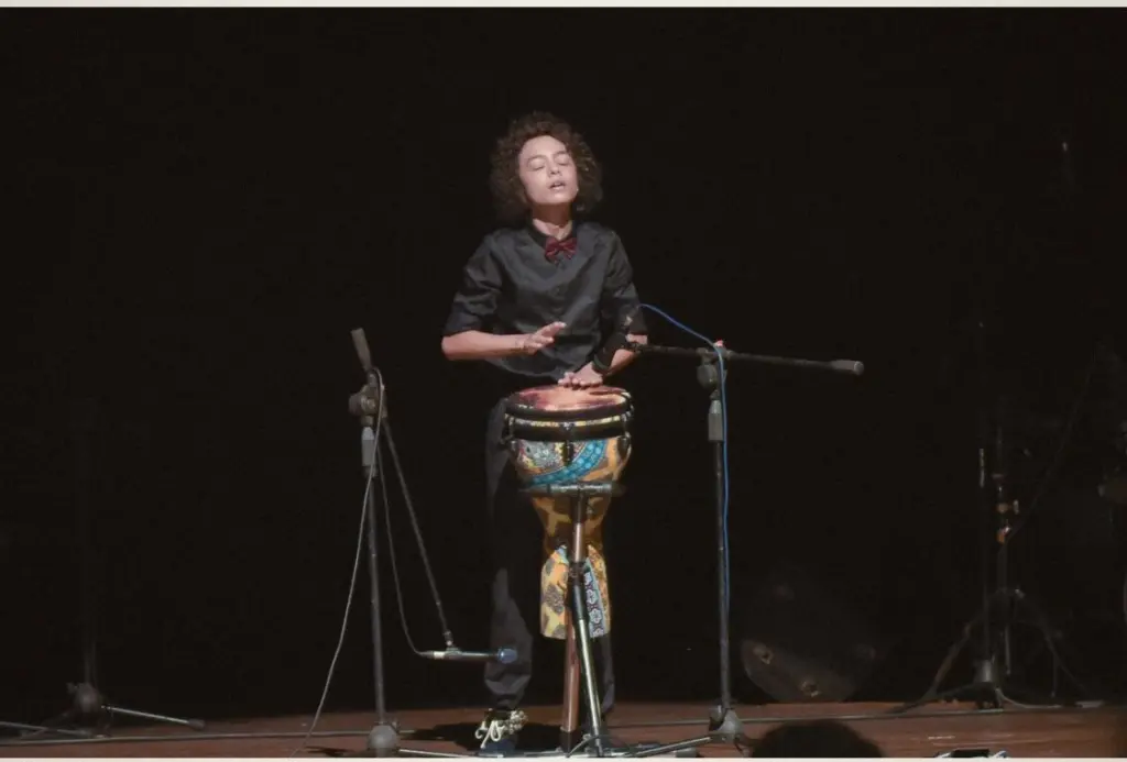 Aurum playing the djembe in black on a dark stage